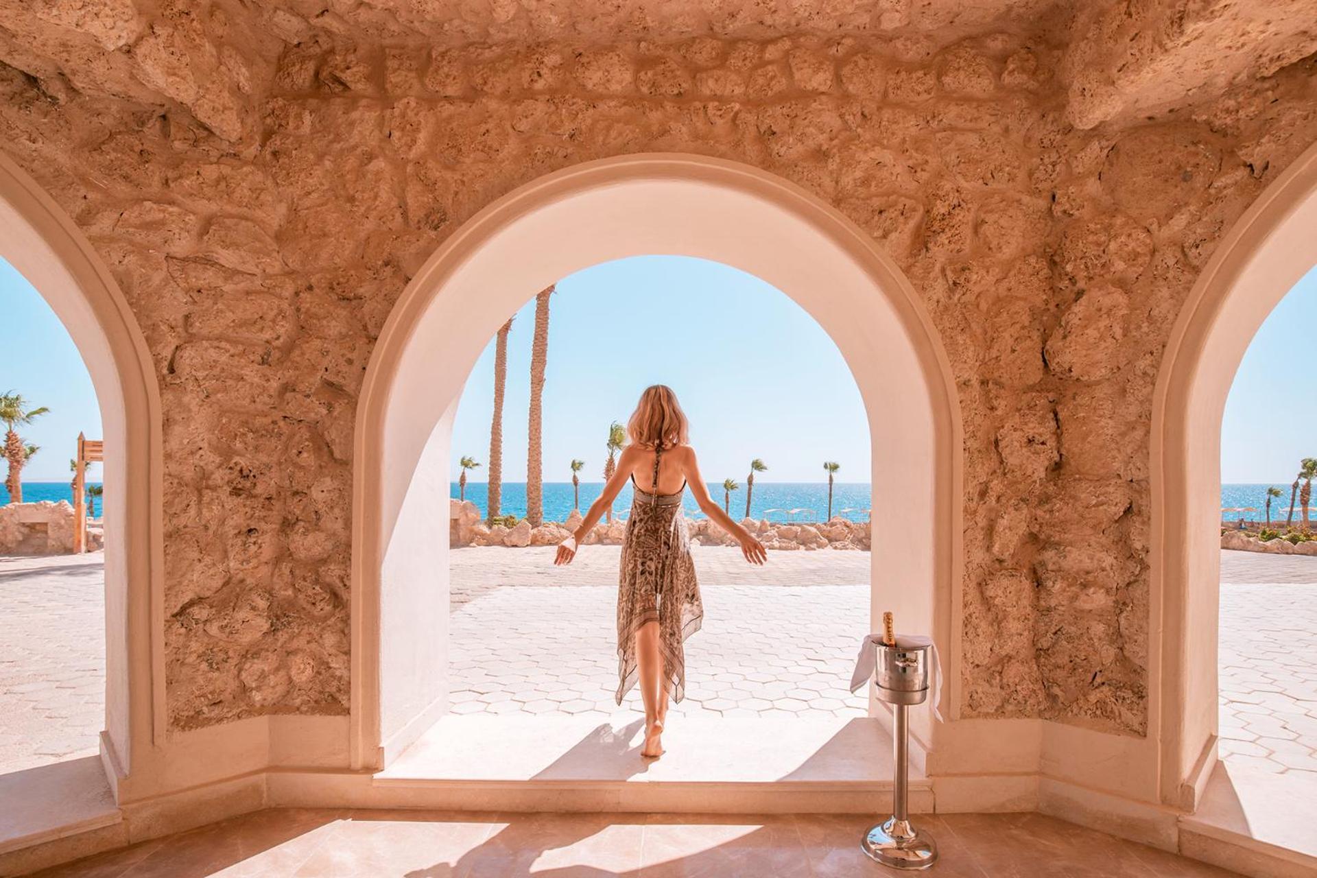 Pickalbatros Citadel Resort Sahl Hasheesh (Adults Only) Хургада Екстер'єр фото The photo depicts a woman standing at an archway that opens up to a scenic view. She is wearing a flowing, patterned dress and appears to be stepping outside. In the background, the vibrant blue sea can be seen, along with palm trees and a clear sky 